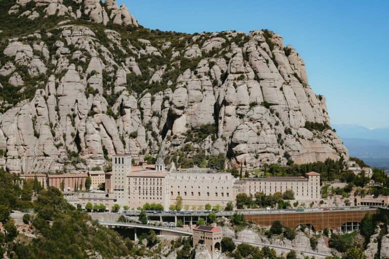 A historic monastery complex nestled against rugged, towering rock formations under a clear blue sky, with modern infrastructure such as a bridge in the foreground.