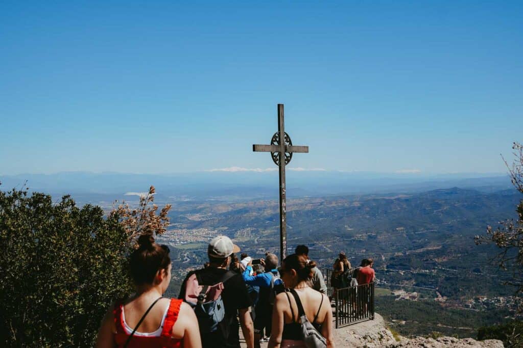 A group of people walk towards a large cross situated on a scenic mountain overlook with a wide view of the landscape and distant mountains, making it a perfect addition to any Montserrat itinerary.