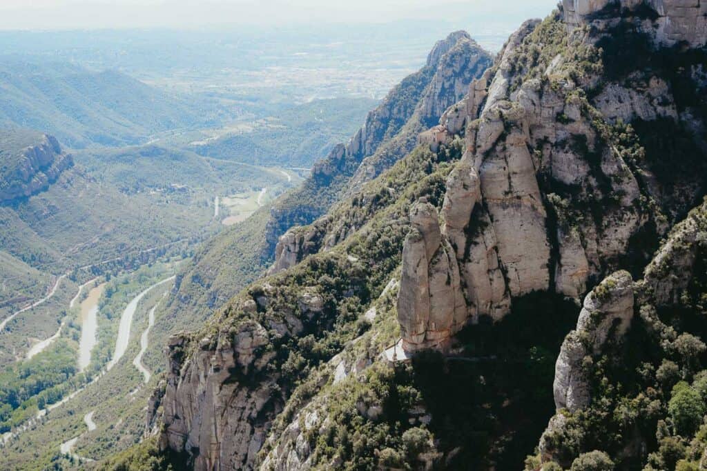 Aerial view of a mountainous landscape featuring rugged cliffs and a winding river valley below, with greenery covering the area and distant rolling hills on a clear day—a scene straight out of a Montserrat itinerary.