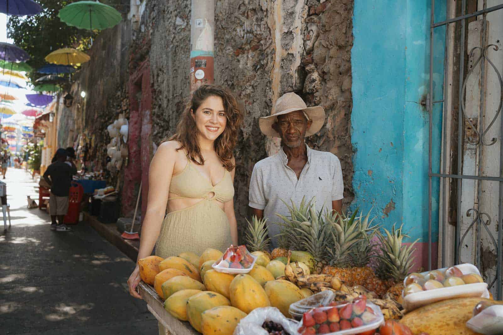 A woman and a man stand beside a fruit stall on a colorful street. The stall displays various fruits like pineapples, bananas, and papayas. Multicolored umbrellas hang overhead, adding to the vibrant scene much like how Lingoda reviews highlight the engaging experience of learning languages online.