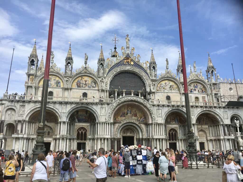 People gather in front of St. Mark's Basilica, an ornate historic church with intricate stonework and mosaics, under a partly cloudy sky, echoing the travel crisis seen in Barcelona overtourism.
