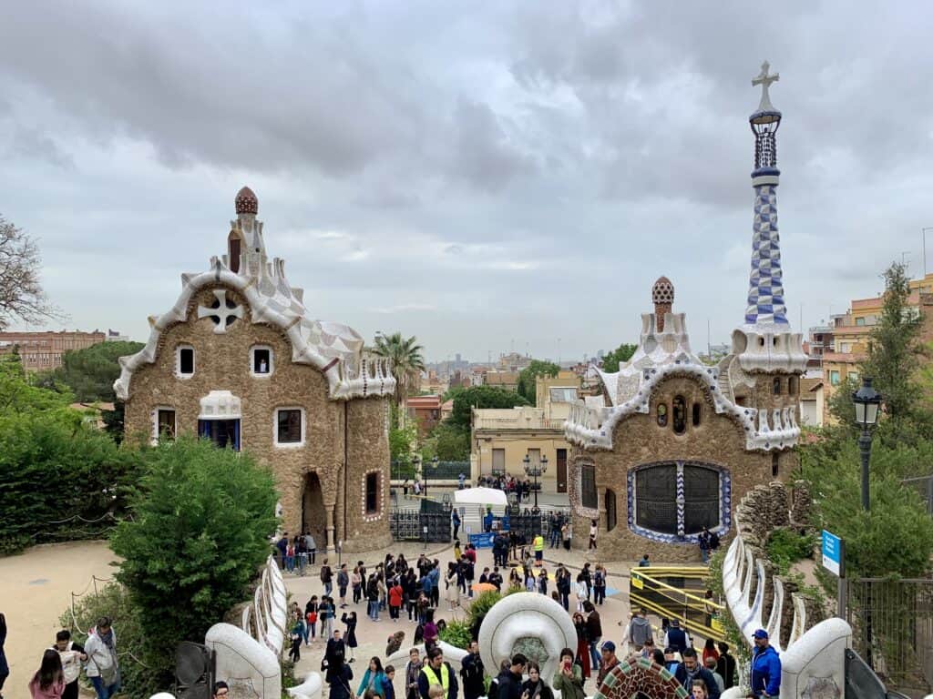 A crowd of people gather in the foreground of two uniquely designed buildings with mosaic roofs in Park Güell, Barcelona, under a cloudy sky, highlighting the city's ongoing struggles with overtourism.