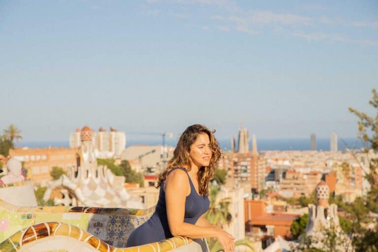 A woman with curly hair leans on a decorative stone railing, overlooking a cityscape with modern and historic buildings under a clear sky, contemplating her 3-day itinerary in Barcelona.