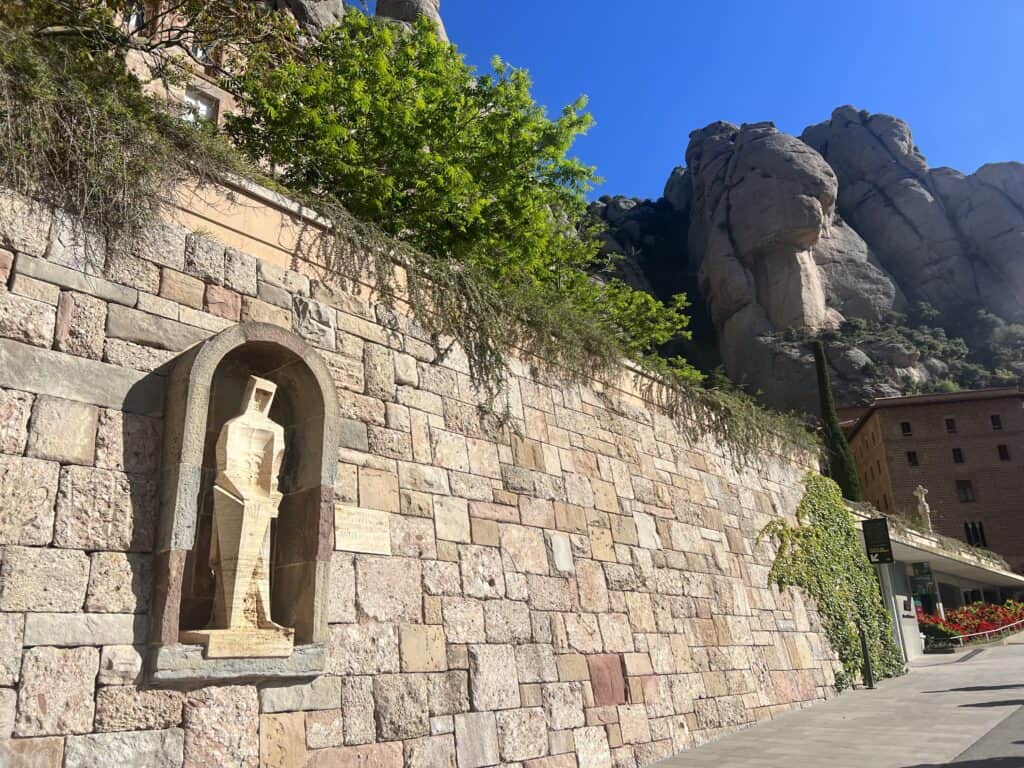 A stone statue is set within a niche on a brick wall with mountains in the background and a building partially visible on the right, resembling a serene stop on any Montserrat itinerary.
