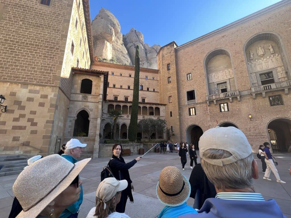 A tour guide speaks to a group of tourists in an outdoor courtyard surrounded by historic brick buildings, with rocky mountains in the background, as they begin their Montserrat itinerary.