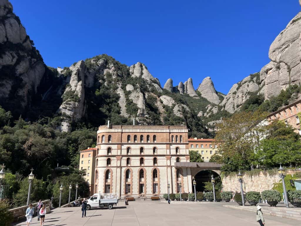 A historical building set against the towering rocky mountains of Montserrat under a clear blue sky, with a few people walking in the foreground, perfect for any itinerary.