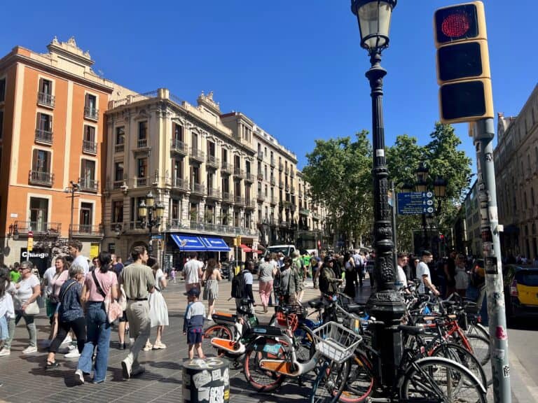 A busy street scene with numerous pedestrians walking, parked bicycles, and a mix of historic and modern buildings under a clear blue sky. A lamppost and traffic light are visible in the foreground, setting the perfect start for your 3 days in Barcelona itinerary.