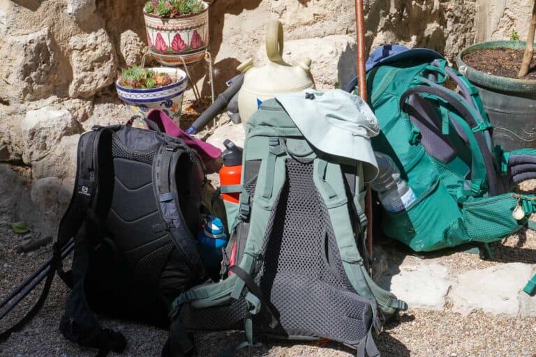Three backpacks rest against a stone wall, surrounded by potted plants and outdoor gear like a water canteen and trekking poles, essential for walking the Camino de Santiago as a digital nomad. A gray shirt is draped over one of the backpacks.