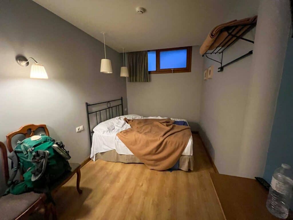 A small bedroom with a metal bedframe, a messy bed with white sheets and a brown blanket, a green backpack on a chair, a wooden floor, and two wall lamps. Bottles are on a wooden table.