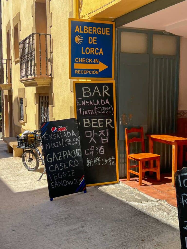 Outdoor entrance of Albergue de Lorca with a yellow sign, two chalkboards advertising menu items like salad, gazpacho, and beer in multiple languages, and a bicycle parked nearby.