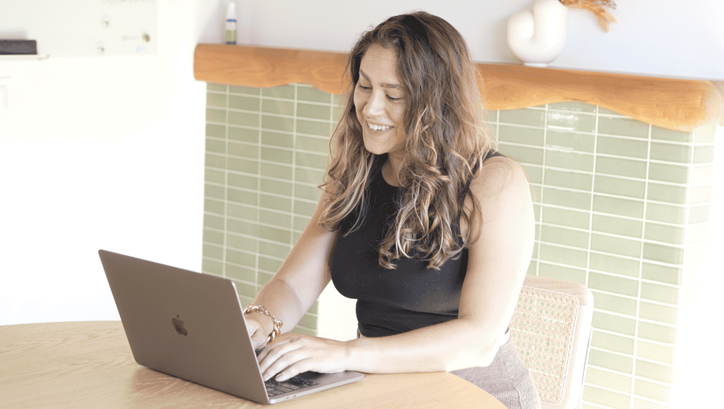 A person with long hair sits at a round wooden table, typing on a laptop and smiling, perhaps working on a language learning plan. Green tile and wood decor are visible in the background.