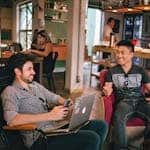 Two men sitting in a café, one holding a smartphone and the other a drink, both smiling. In the background, other patrons and staff are visible. The scene captures the essence of walking the Camino de Santiago as a digital nomad, blending work with adventure seamlessly.