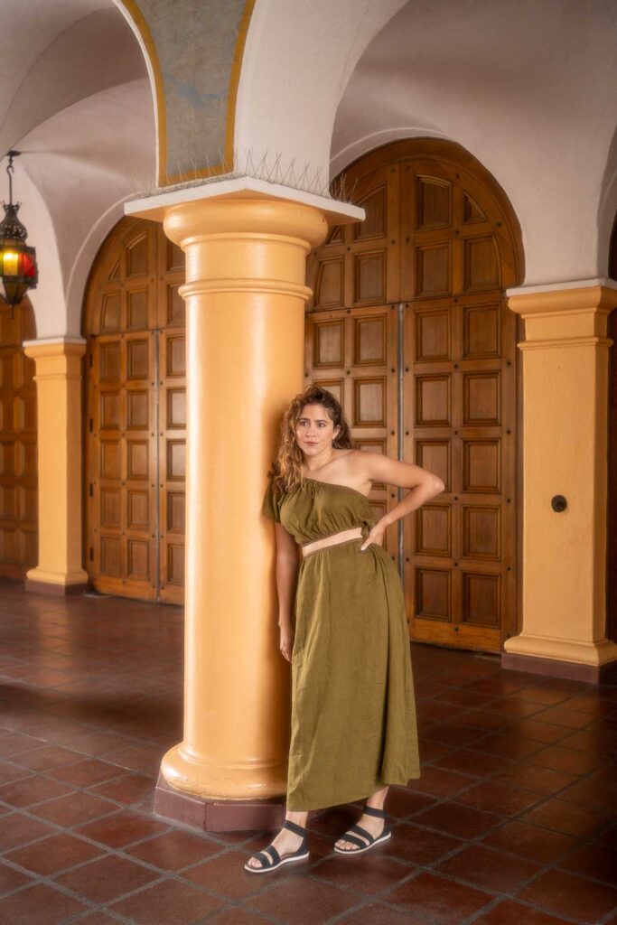 A woman in a green one-shoulder dress leans against a yellow column in a hallway with arched ceilings and wooden doors, showcasing the perfect inspiration for what to wear in Los Angeles.