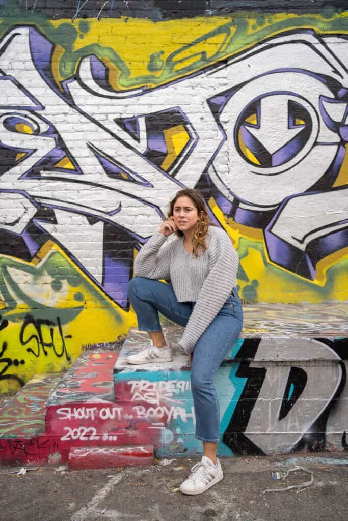 A person in a gray sweater and blue jeans, a perfect casual choice for what to wear in Los Angeles, poses with one leg up on a step in front of a colorful graffiti wall.