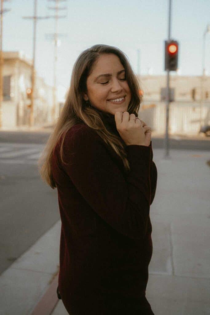 A woman with long hair stands on a city sidewalk, smiling with her eyes closed and hands near her face. She wears a dark long-sleeved top, perfect for any local wondering what to wear in Los Angeles. A traffic light is visible in the background.