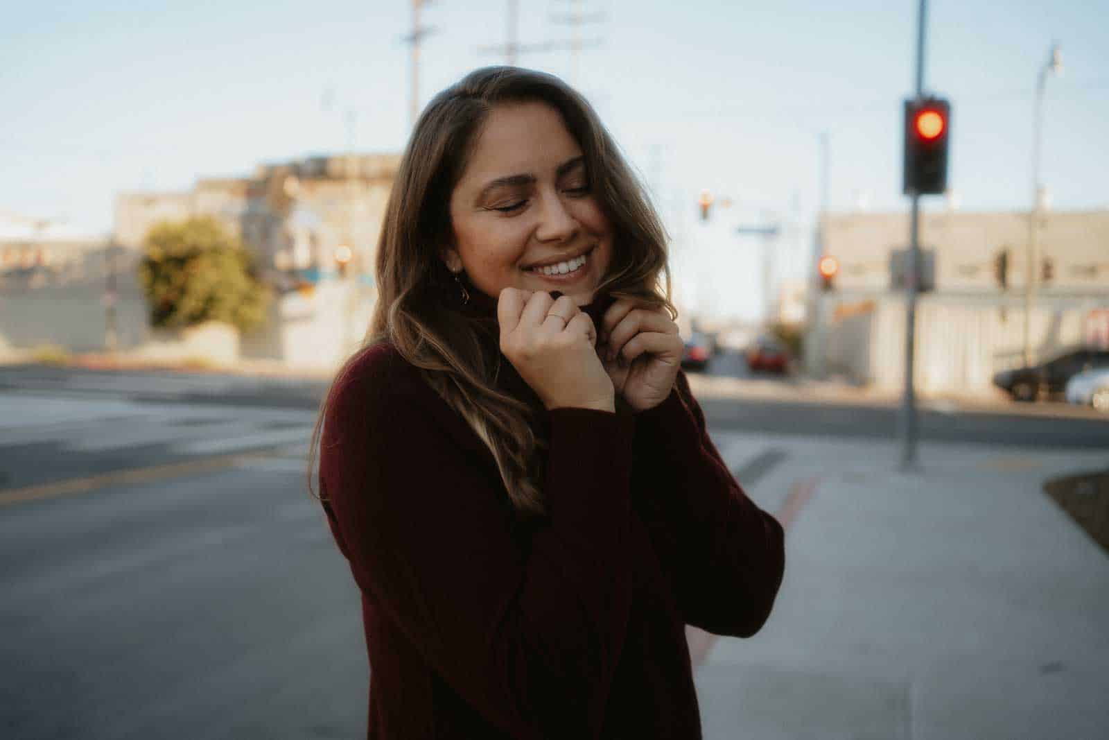 A person stands on a city street, smiling with eyes closed, holding the collar of their dark sweater. Traffic lights and buildings are visible in the background, offering a glimpse into what to wear in Los Angeles regardless of every season.