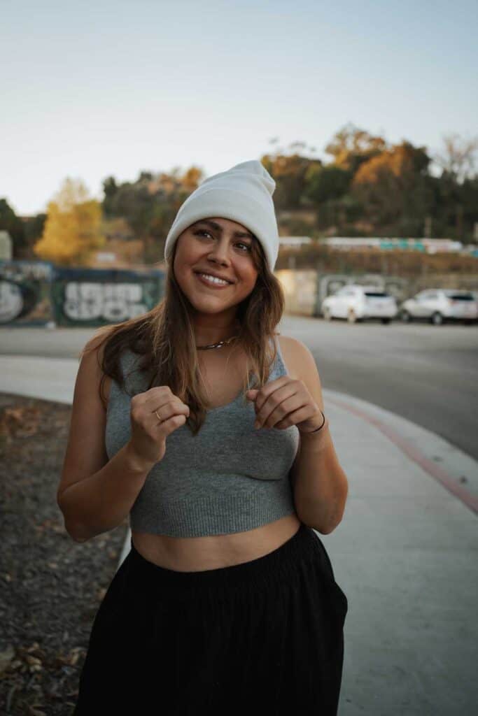 A woman wearing a white beanie, gray tank top, and black pants stands outside on a sidewalk in Los Angeles, smiling. Trees and parked cars are in the background, providing a snapshot of what to wear in Los Angeles for a casual day out.