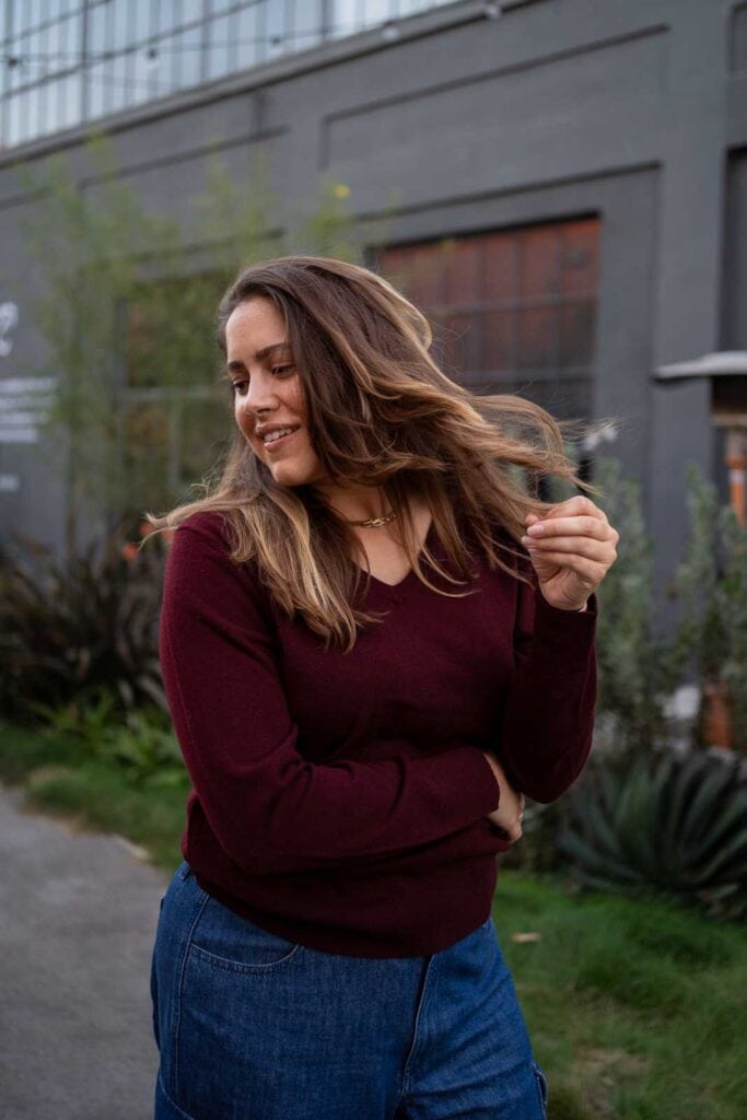 A woman with long brown hair, dressed in a maroon sweater and blue jeans—perfect examples of what to wear in Los Angeles—stands outdoors, lightly holding her hair with one hand and smiling slightly. She's in front of a modern building with plants nearby.