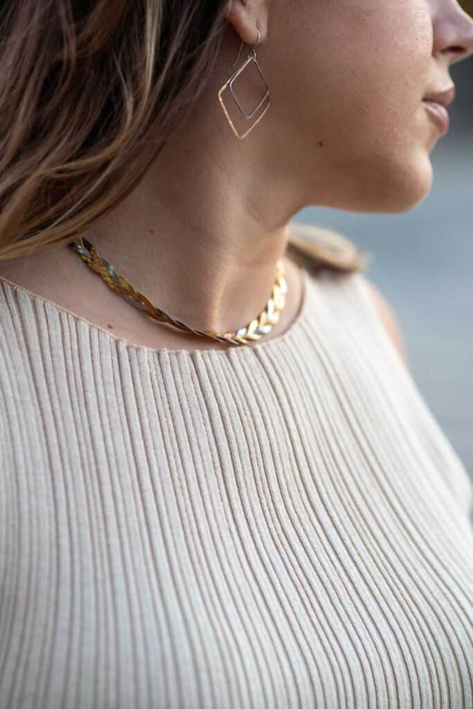 Close-up of a person wearing a gold leafy necklace, a thin geometric earring, and a sleeveless, ribbed, beige top. Their face is partially visible, turned slightly to the side. A true Los Angeles fashion guide on what to wear this season.