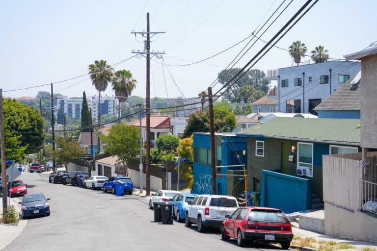 Driving in Los Angeles, you'll often find yourself on a suburban street with parked cars, houses, utility poles, and palm trees on a clear day. Tips from a local: always keep an eye out for hidden driveways and ensure pedestrians have ample space to navigate the serene neighborhood.
