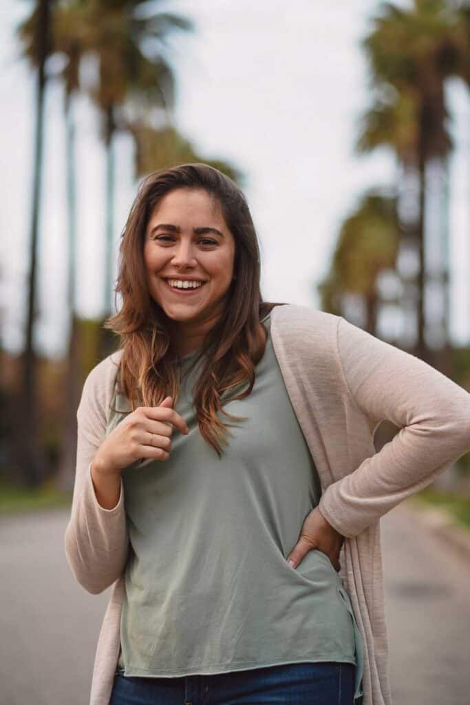 A person with long brown hair, wearing a light green shirt and beige cardigan, smiles while standing outdoors with palm trees in the background, showcasing a perfect example of what to wear in Los Angeles.