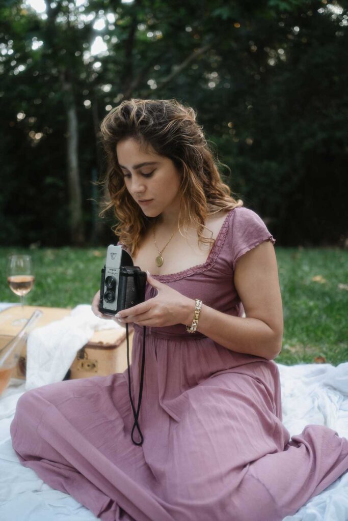 A woman in a purple dress sits on a picnic blanket in a park, looking at a vintage camera in her hands. A glass of wine and picnic items are beside her. Trees are visible in the background, showcasing an ideal choice for what to wear in Los Angeles during a relaxed afternoon outing.