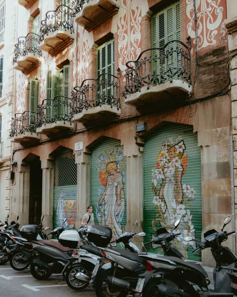 Street scene in Barcelona in the summer, with parked scooters in front of a building adorned with large decorative murals and ornate balconies.