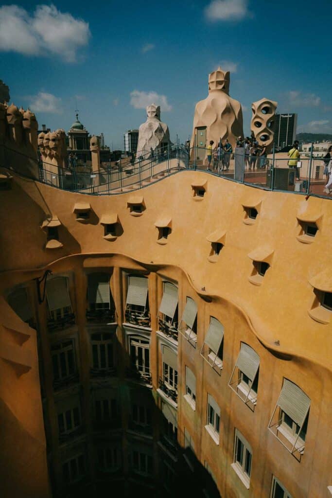 On a sunny summer day in Barcelona, the rooftop of a building with wavy yellow facades and unique sculptural chimneys comes alive. Visitors wander, soaking in the beautiful views beneath the blue sky dotted with scattered clouds.