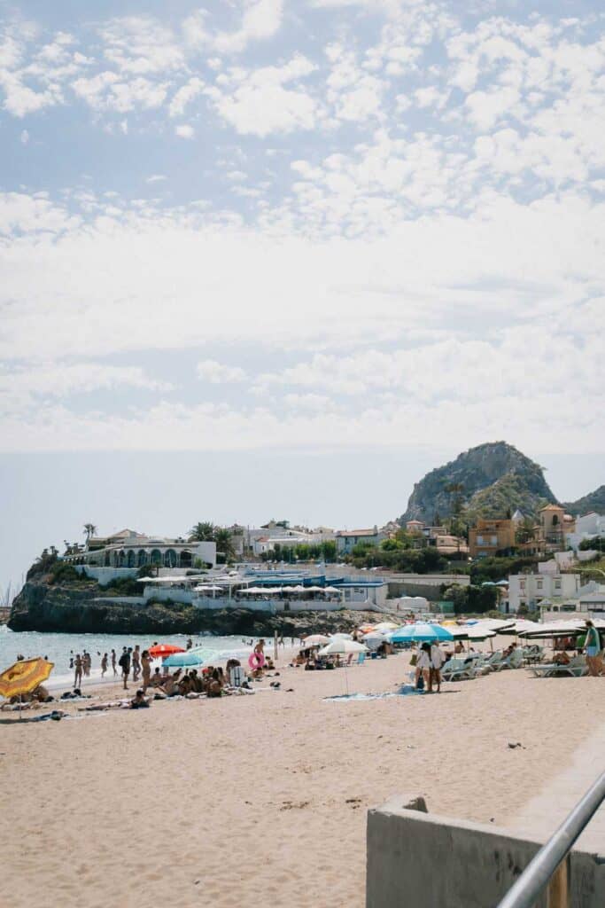 The beach scene captures the essence of Barcelona in the Summer, with people relaxing under umbrellas and basking in the sun's warmth. In the background, charming houses and a rocky hill add to the picturesque view.
