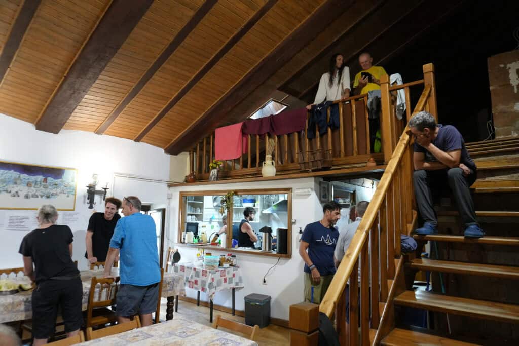 People interacting in a cozy wooden room with an open kitchen and a loft area reminiscent of sleeping on the Camino. Some are sitting, others are standing or talking on the stairs. The warm lighting enhances the relaxed atmosphere.