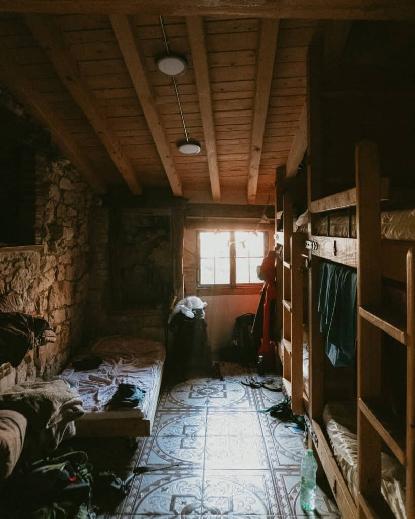 A rustic, dimly lit room with wooden bunk beds awaits weary travelers on the Camino de Santiago. Stone walls and a tiled floor echo serenity, while a small window offers soft light to ensure a good night's rest. Clothing and items are scattered on the beds and floor in relaxed disarray.