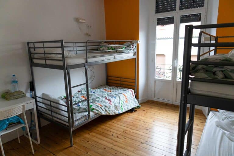 A room with two metal bunk beds, one set unmade, reminiscent of nights spent sleeping on the Camino de Santiago. A small desk with a water bottle sits to the side. A window with shutters frames the background, and the walls are painted in soothing orange and white tones.