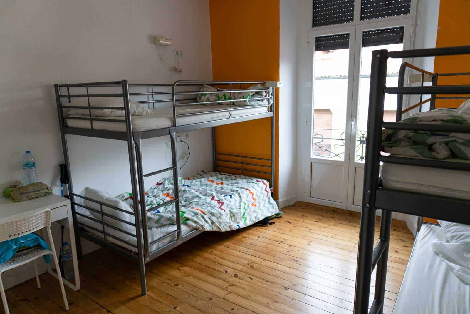 A room with two metal bunk beds, one set unmade, reminiscent of nights spent sleeping on the Camino de Santiago. A small desk with a water bottle sits to the side. A window with shutters frames the background, and the walls are painted in soothing orange and white tones.