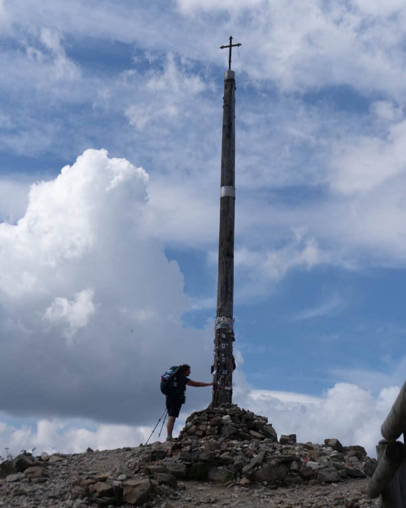 A weary traveler sleeping on the Camino awakens to a somber sky and approaches a tall wooden cross on a stone mound, reaching out with reverence.