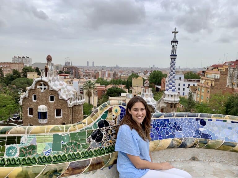 A person sits on a colorful mosaic bench in Barcelona, enjoying a cityscape view even on a cloudy day.
