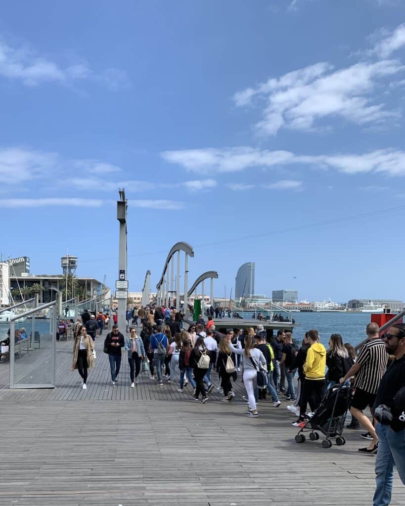 People strolling on a wooden pedestrian bridge with modern arch structures over water, the Barcelona skyline visible in the background under a partly cloudy summer sky.