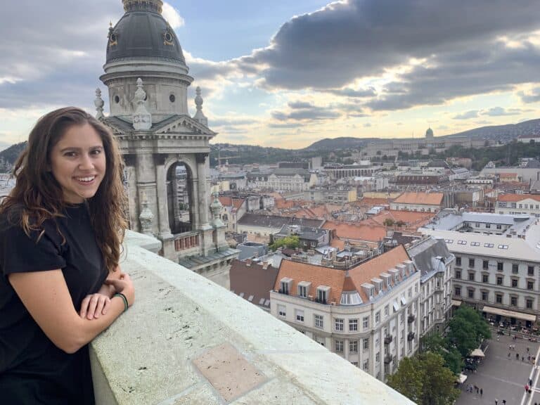 A woman leans on a stone ledge, pondering digital nomad tips, as she gazes out over a cityscape filled with historic buildings beneath a cloudy sky.