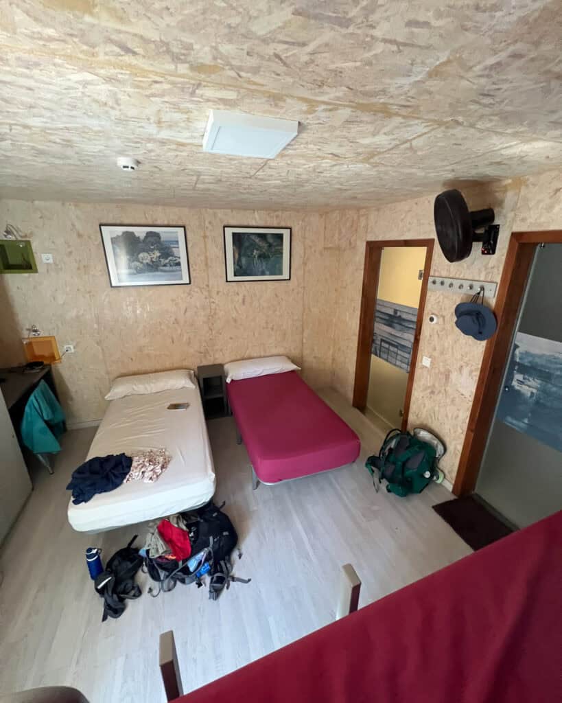 A cozy room perfect for a night's rest after a day on the Camino de Santiago, featuring two single beds, a wall-mounted TV, and a door. Clothes and backpacks scatter the floor as two framed pictures add warmth to the walls.