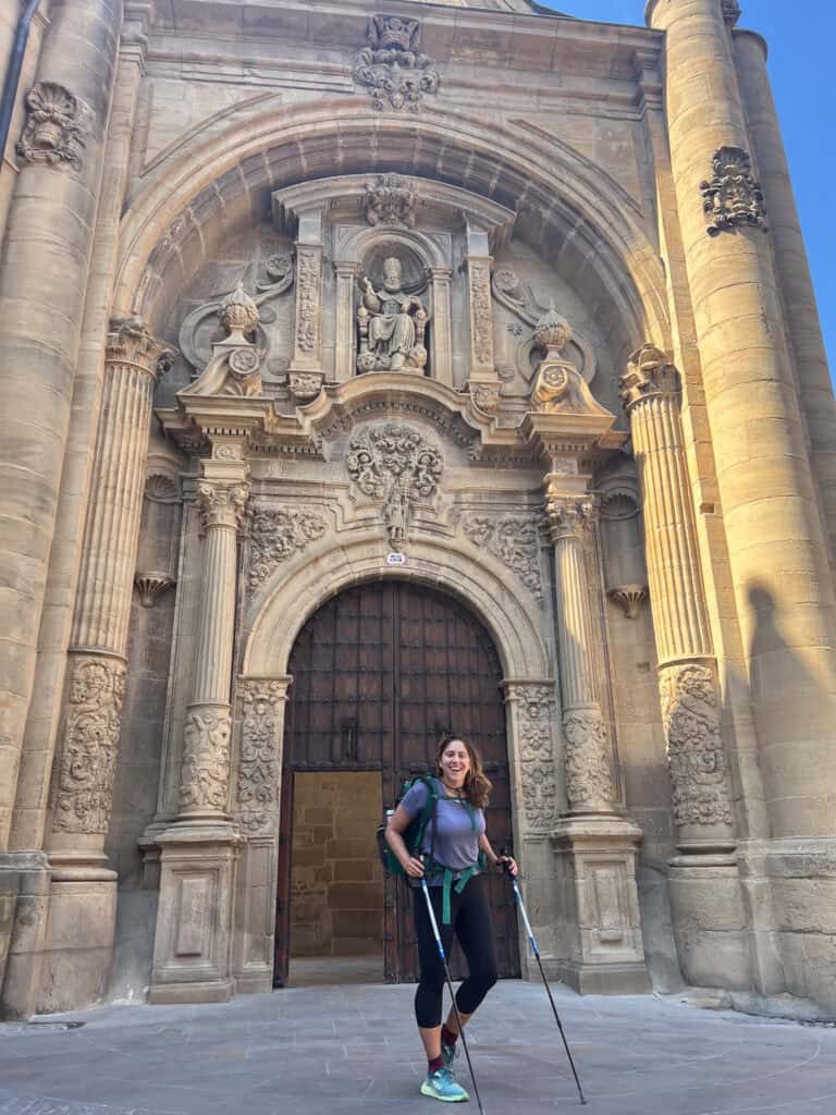 A person with hiking gear stands before an ornate stone church facade, detailed carvings catching the light, reminiscent of tales from nights spent sleeping on the Camino. The arched wooden doors beckon travelers to pause and reflect.