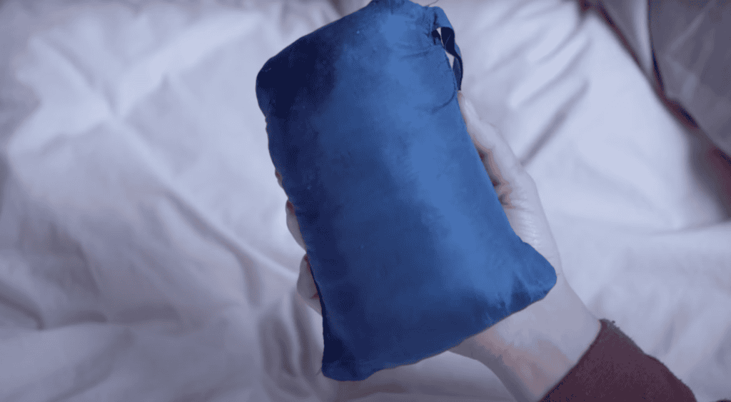 A hand holding a small, blue, drawstring travel pillow on a white bedspread, perfect for those nights sleeping on the Camino.
