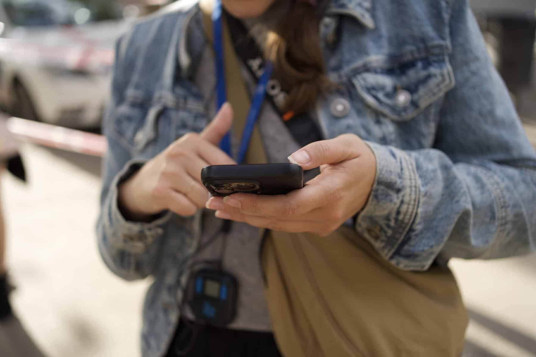 A person wearing a denim jacket uses a smartphone outdoors, with a lanyard and small device hanging from their neck—perfectly equipped with an eSIM for Europe for seamless connectivity.