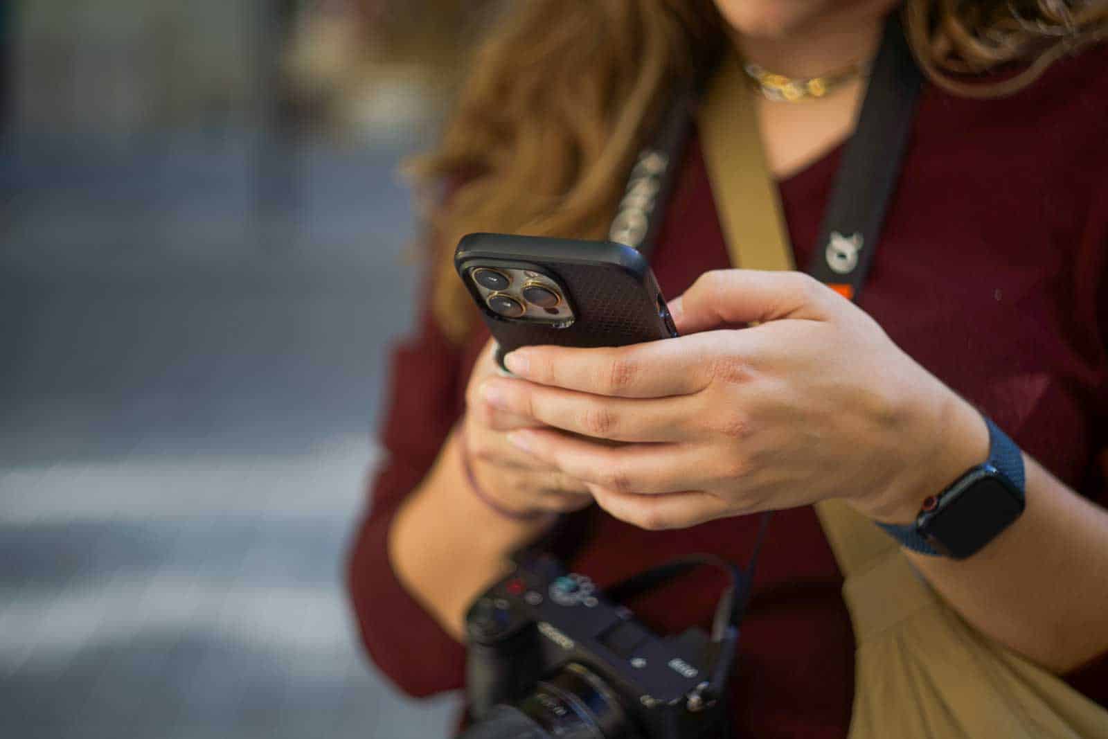 Person holding a smartphone, looking at the screen. A camera is hanging from their neck.