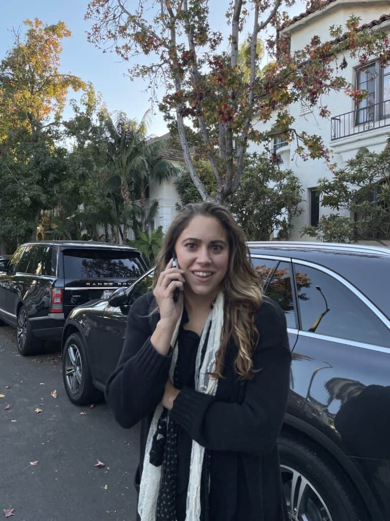 A person is standing on a street, talking on a phone about esims for digital nomads. They are wearing a black jacket and a scarf. Parked cars line the street, and trees are in the background.