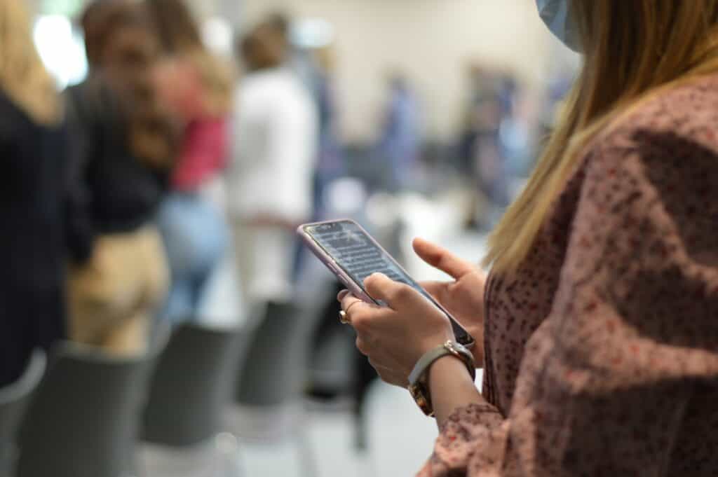 A person wearing a mask is holding a smartphone, likely exploring esims for digital nomads, with blurred people and chairs in the background.