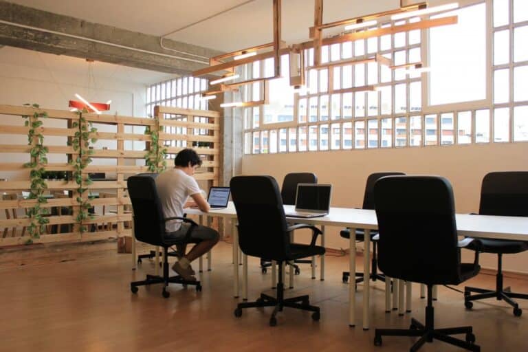 A person sits at a long table with multiple chairs in a spacious, well-lit office, reminiscent of Barcelona coworking spaces. Laptops are scattered on the table, while a wooden partition adorned with plants forms the backdrop.