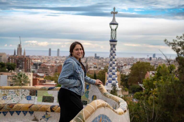 A person in a denim jacket leans on a mosaic railing, overlooking a cityscape with distinctive towers and a cloudy sky.