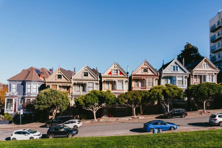 A row of colorful Victorian houses with cars parked in front and trees lining the street, set under a clear blue sky—an iconic scene you'll encounter during your one day in San Francisco.