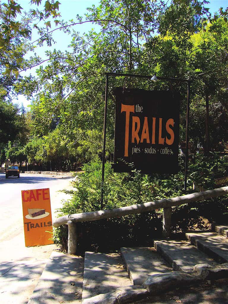 Outdoor signs for The Trails Cafe, near the Griffith Observatory hike, are posted beside a tree-lined road and stone steps, advertising pies, sodas, and coffee.