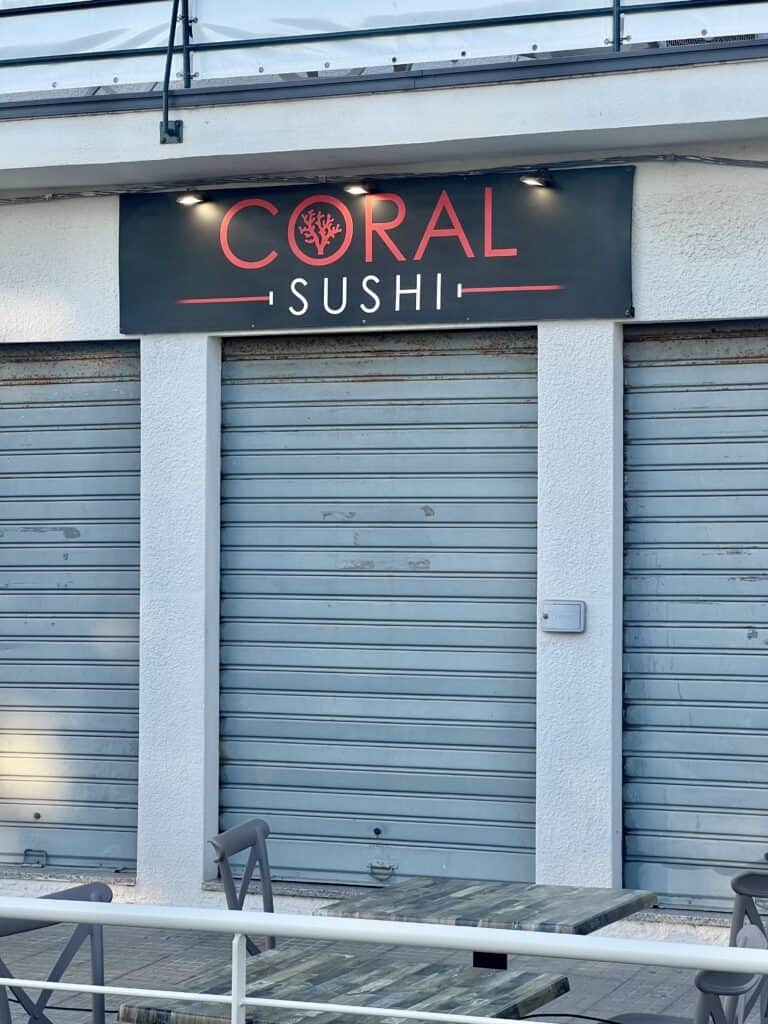 A closed restaurant with a sign reading "Coral Sushi" above metal shutters, with outdoor tables and chairs in front—once a popular spot among the best restaurants Alghero has to offer.