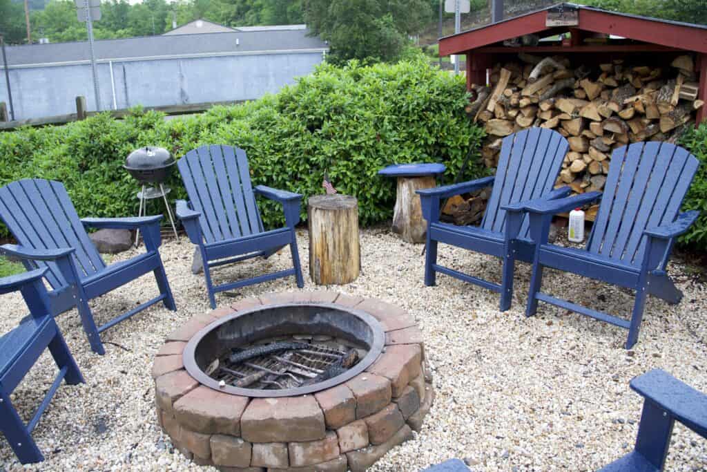 Five blue Adirondack chairs surround a stone fire pit on gravel, with a woodpile and metal grill visible in the background—a cozy setting often praised in Western Front Hotel reviews.